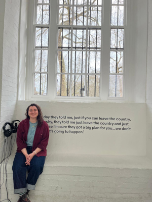 Millie Shafiee sits on the left of a window ledge in a white walled gallery space, the ICA. There is writing underneath the window in black that reads ‘One day they told me, just if you can leave the country. I asked why, they told me just leave the country and just go because I’m sure they got a big plan for you…we don’t know what’s going to happen’. There are headphones attached to the wall on the left of the installation.