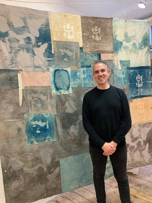 A photo of artist JC Candanedo standing in front of one of his textile pieces made of a patchwork of cyanotype prints on blue and brown dyed fabric. He is smiling, wearing black jumper with black trousers that complement the artistic setting.