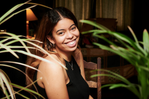 A mixed-race girl is smiling while wearing a black top. She is sat on a chair surrounded by green leaves.